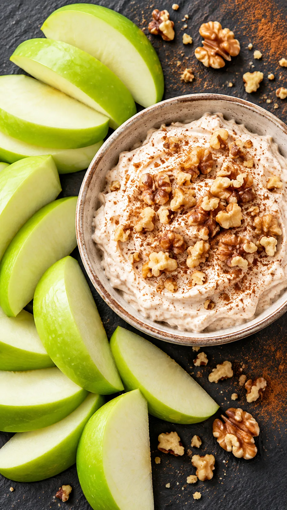 Overhead view of crisp green apple slices placed next to a small ceramic bowl filled with whipped cinnamon cottage cheese dip, sprinkled with crushed walnuts on a slate background