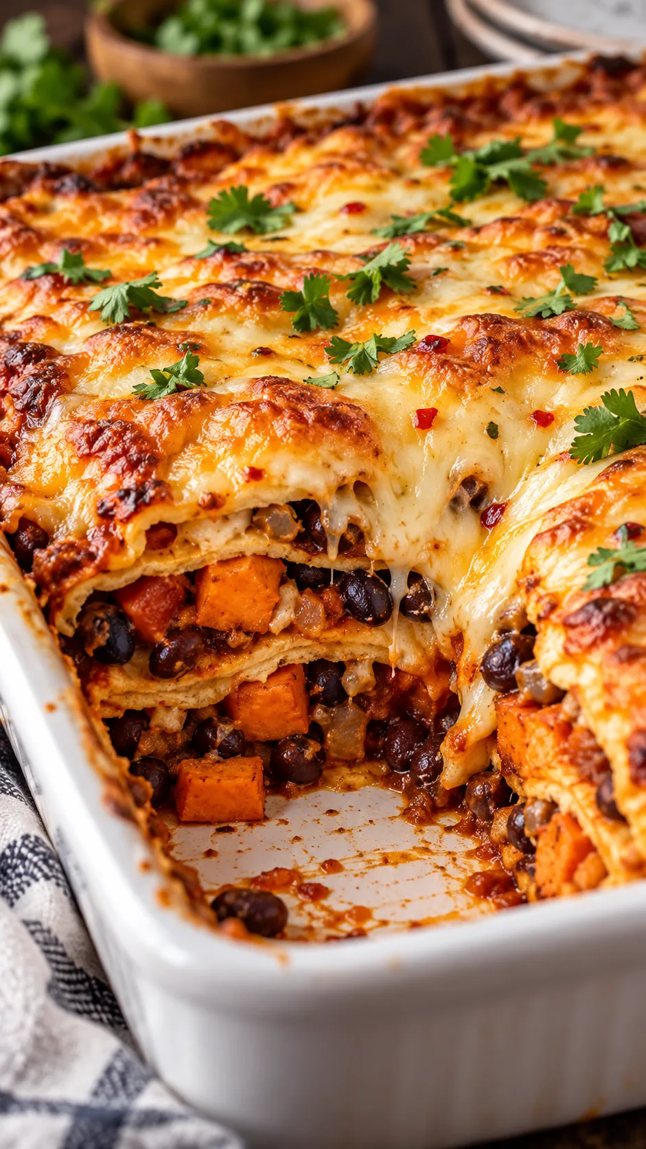 A baked square enchilada casserole cut into, showing layers of corn tortillas, black beans, sweet potatoes, and melted bubbling golden brown pepper jack cheese on top