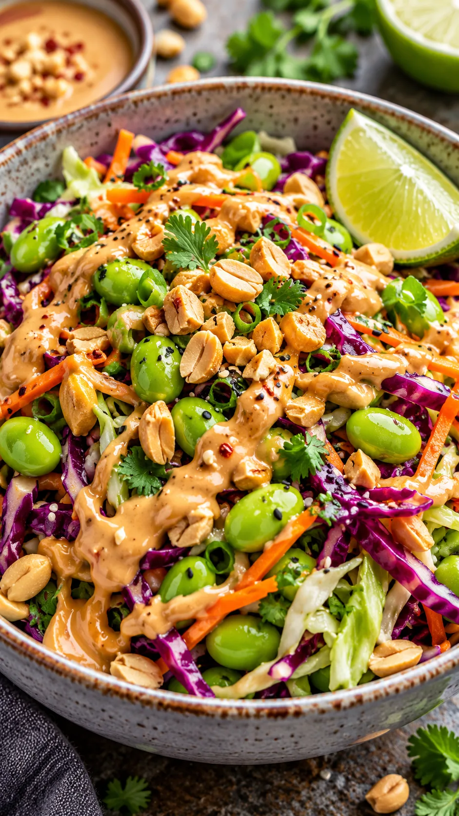 Close up shot of a vibrant colorful Asian-inspired edamame cabbage salad bowl with thick peanut dressing swirl, roasted peanuts, and lime wedge