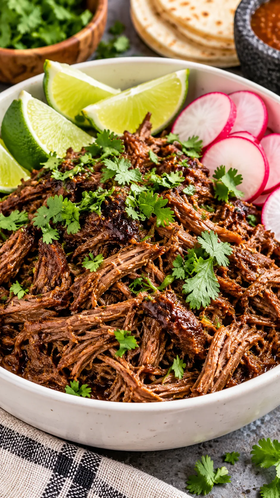 Tender shredded barbacoa beef piled high in a white bowl with fresh lime wedges, fresh chopped cilantro, and sliced red radishes