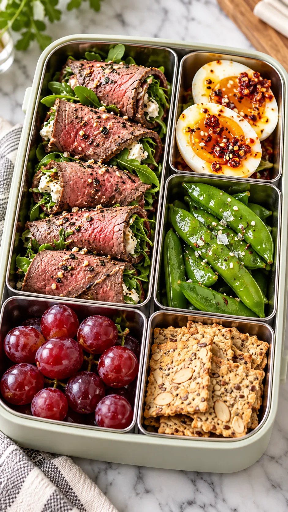 A high-end multi-compartment bento lunch box containing sliced roast beef roll-ups, soft-boiled eggs with chili crunch, crunchy sugar snap peas, red grapes, and a compartment of almond flour crackers