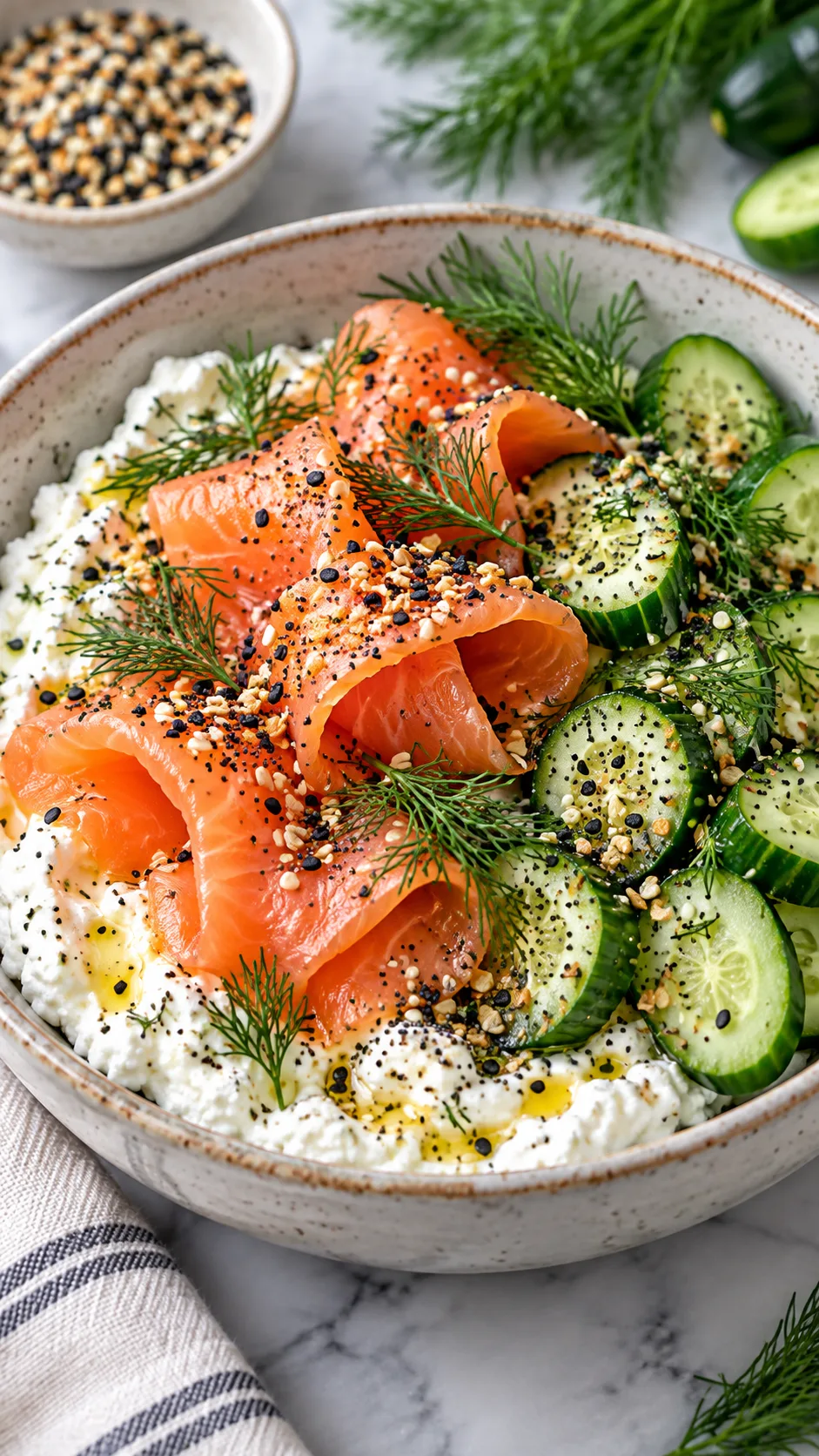 A delicious meal prep bowl containing whipped cottage cheese topped with folded cold-smoked salmon ribbons, sliced baby cucumbers, fresh green dill, and a sprinkle of everything bagel seasoning.