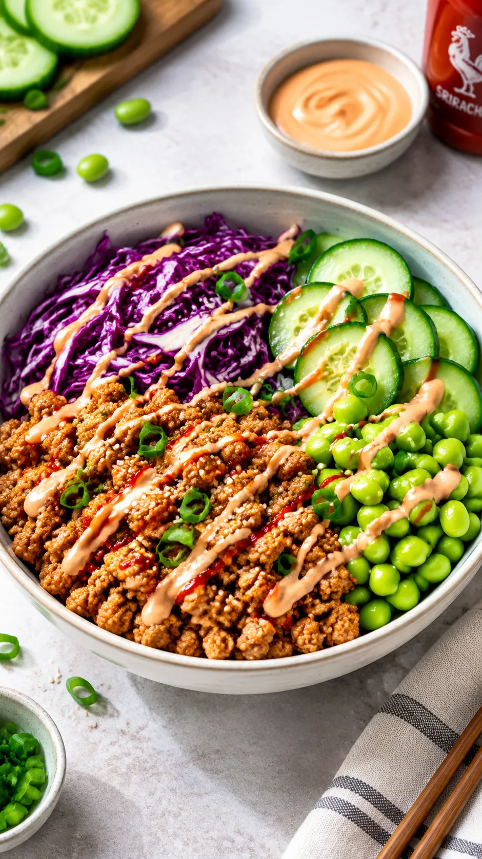 Clean, modern high protein bowl filled with browned ground turkey, shredded red cabbage, sliced cucumbers, edamame, and a drizzle of sriracha mayo